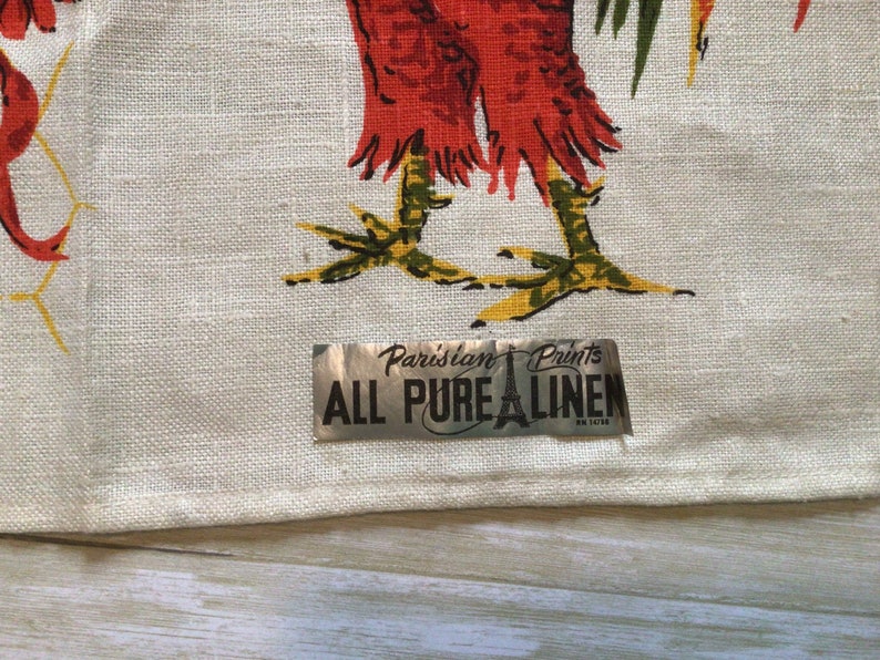 May include: A white linen tea towel with a rooster design and a silver label that reads "Parisian Prints All Pure Linen".