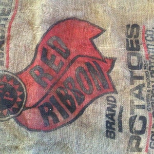 May include: Close-up of a burlap potato sack with the words "RED RIBBON BRAND POTATOES" printed in black and red. The sack has a worn, vintage appearance, with visible texture and faded ink.