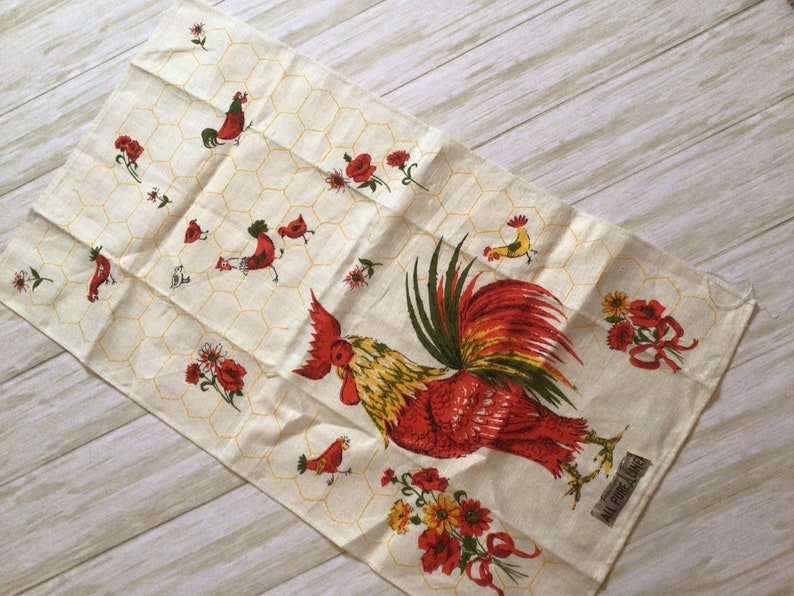 May include: A white tea towel with a red rooster and yellow flowers on a honeycomb pattern. The text "All Purpose Linen" is printed in the bottom right corner.