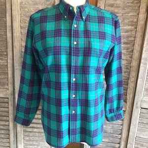 May include: A long-sleeve button-down shirt featuring a plaid pattern in shades of teal green and dark purple. The shirt has a pointed collar, a front button placket with light-colored buttons, and a single chest pocket on the left side.
