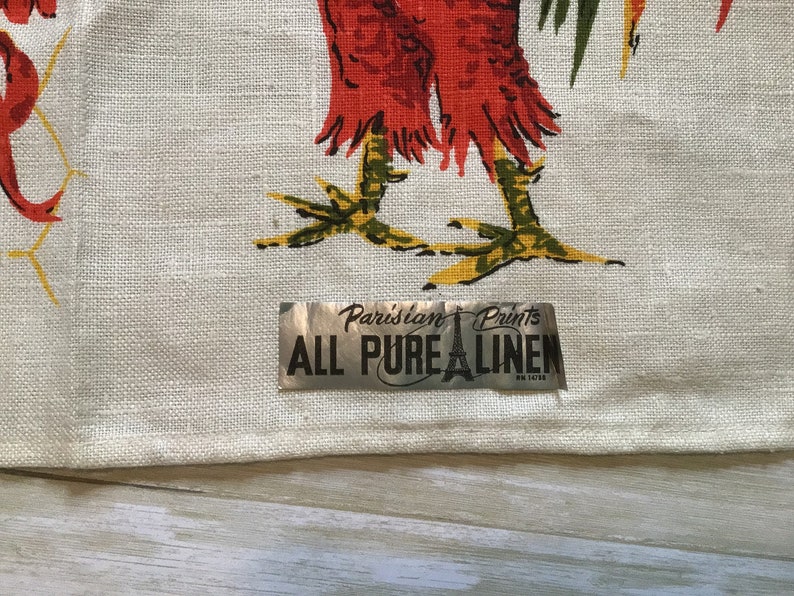 May include: A white linen tea towel with a rooster design and a silver label that reads "Parisian Prints All Pure Linen".