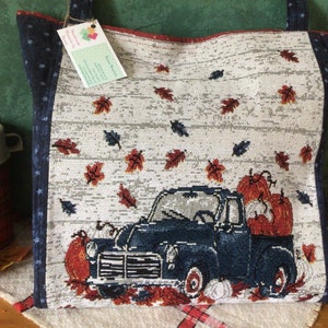 Pu&ograve; includere: Una borsa tote bianca e blu con un camioncino blu che trasporta zucche e foglie autunnali. Il camioncino &egrave; su uno sfondo bianco con un motivo a venature di legno. La borsa ha un motivo a stelle blu e bianco sui lati.