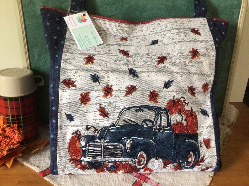 Pu&ograve; includere: Una borsa tote bianca e blu con un camioncino blu che trasporta zucche e foglie autunnali. Il camioncino &egrave; su uno sfondo bianco con un motivo a venature di legno. La borsa ha un motivo a stelle blu e bianco sui lati.