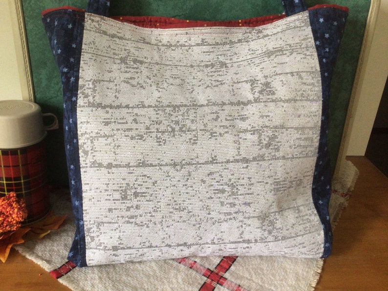 Pu&ograve; includere: Una borsa tote bianca con un motivo a stelle blu e bianche sui lati. La borsa &egrave; realizzata in tessuto intrecciato e ha una striscia rossa e bianca in alto.