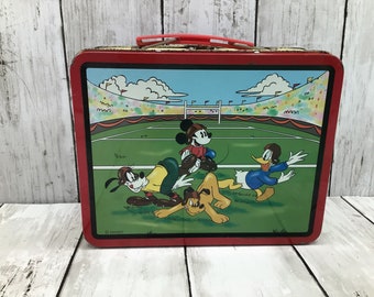 May include: Red metal lunchbox featuring a cartoon image of Mickey Mouse, Goofy, and Donald Duck playing a game of football on a green field. The lunchbox has a handle and the text "© DISNEY" is visible at the bottom.