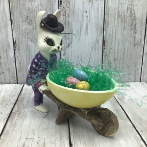 May include: A ceramic Easter bunny figurine wearing a black hat and purple plaid outfit, pushing a yellow wheelbarrow filled with green Easter grass and decorated eggs. The wheelbarrow has a wooden frame.