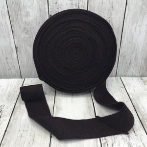 Peut inclure: Un rouleau de ruban en tissu de couleur violet fonc&eacute;, enroul&eacute; en cercle, avec une longueur de ruban d&eacute;roul&eacute;e sur une surface en bois blanc.