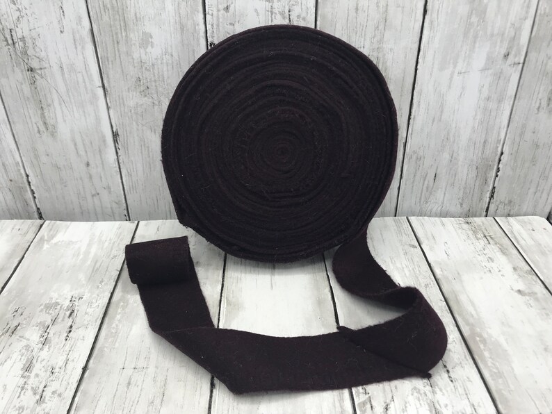Peut inclure: Un rouleau de ruban en tissu de couleur violet fonc&eacute;, enroul&eacute; en cercle, avec une longueur de ruban d&eacute;roul&eacute;e sur une surface en bois blanc.