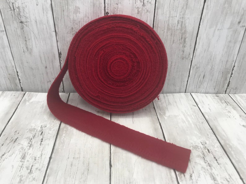 Peut inclure: Un rouleau de galon en tissu rouge, d'environ 3,8 cm de large, est d&eacute;roul&eacute; sur un fond en bois blanc. Le tissu est de couleur rouge unie et a une surface l&eacute;g&egrave;rement textur&eacute;e.