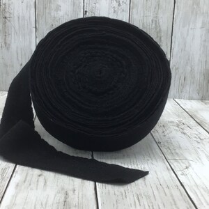 Peut inclure: Un rouleau de tissu en feutre noir, d'environ 2,5 cm de large, est pr&eacute;sent&eacute; sur une surface en bois blanc. Le feutre est enroul&eacute; serr&eacute; et l'extr&eacute;mit&eacute; du rouleau est d&eacute;roul&eacute;e.