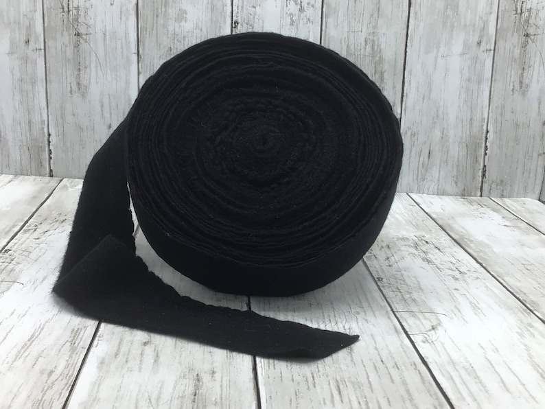 Peut inclure: Un rouleau de tissu en feutre noir, d'environ 2,5 cm de large, est pr&eacute;sent&eacute; sur une surface en bois blanc. Le feutre est enroul&eacute; serr&eacute; et l'extr&eacute;mit&eacute; du rouleau est d&eacute;roul&eacute;e.