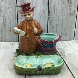 May include: A ceramic figurine of a man wearing a brown coat and a top hat, holding a small plate. The figurine is standing on a green and blue base with two small bowls. The base has the number 8621 printed on it.