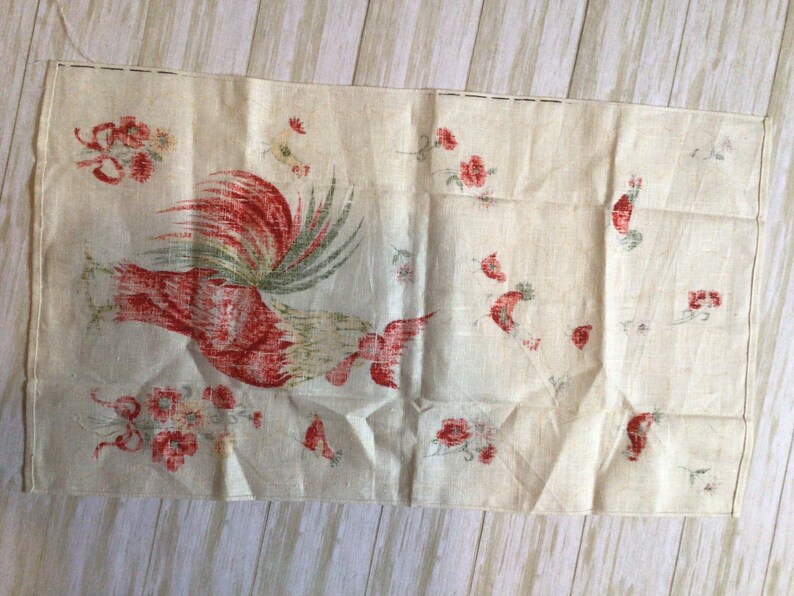 May include: A vintage linen tea towel with a red rooster and floral design. The rooster is facing right and has a green tail. The flowers are red and white.