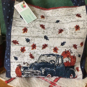 Pu&ograve; includere: Una borsa tote blu e bianca a tema autunnale. La borsa presenta un camioncino blu con zucche nel cassone e una spolverata di foglie rosse e arancioni. La borsa ha un motivo a stelle blu e bianche sui lati.