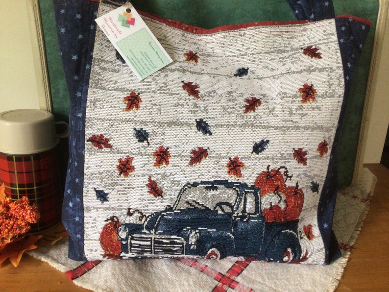 Pu&ograve; includere: Una borsa tote blu e bianca a tema autunnale. La borsa presenta un camioncino blu con zucche nel cassone e una spolverata di foglie rosse e arancioni. La borsa ha un motivo a stelle blu e bianche sui lati.
