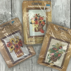 May include: Three vintage Caron Hallmark Design Collection needlepoint kits featuring floral arrangements in baskets. The kits are packaged in plastic and feature a brown wooden frame with the Caron Hallmark Design Collection logo.
