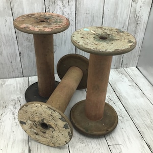 May include: Three vintage wooden spools with distressed paint. The spools have a round base and top, connected by a central column. The paint on the tops is peeling, revealing layers of color. The spools are arranged on a white wooden surface.