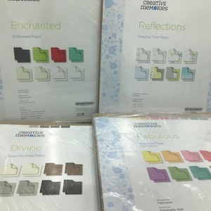 May include: Four packages of Creative Memories paper, each with a different design and title: Enchanted, Reflections, Divine, and Fabulous. The packages are white with a blue and white floral pattern.