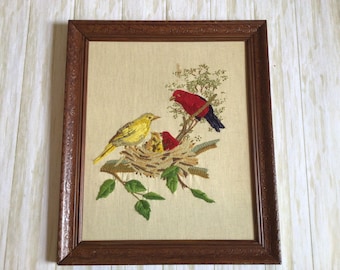 May include: Framed embroidery depicting a family of birds. A yellow bird sits on a nest with two red baby birds. A red bird with a black tail sits on a branch above the nest.