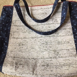 Pu&ograve; includere: Una borsa tote bianca con rifiniture in tessuto blu a stelle. La borsa ha due manici lunghi e una fodera rossa.