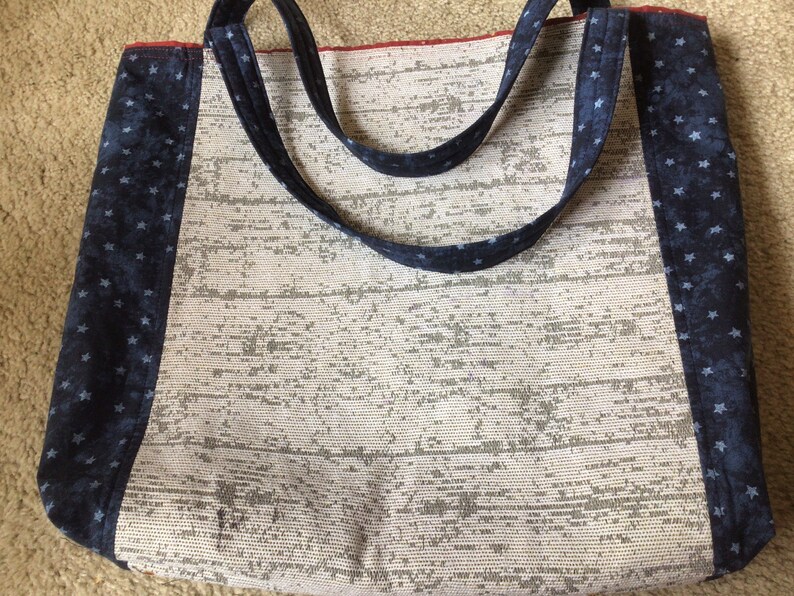 Pu&ograve; includere: Una borsa tote bianca con rifiniture in tessuto blu a stelle. La borsa ha due manici lunghi e una fodera rossa.