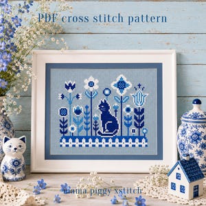 Op de afbeelding: Een PDF-kruissteekpatroon met een donkerblauwe kat die in een tuin van gestileerde blauwe en witte bloemen en een hek zit. Het ontwerp wordt getoond in een witte lijst met een blauwe passe-partout, met de tekst PDF cross stitch pattern erboven.
