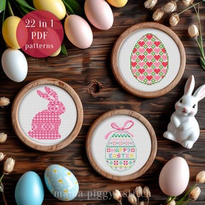 Puede incluir: Patrones de punto de cruz enmarcados en madera con temas de Pascua. Los diseños incluyen un conejito rosa, un huevo con corazones y un huevo con el texto "Happy Easter". La imagen también presenta huevos decorativos y un conejito de cerámica sobre una superficie de madera.
