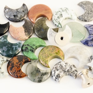 May include: A collection of 17 crescent moon shaped polished stones in various colors and patterns, including black, green, brown, white, and blue. The stones are arranged on a white surface.