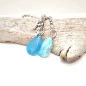 May include: Two blue agate teardrop pendants hang from a silver chain. The chain is made of small silver beads. The pendants are attached to the chain with silver rings.