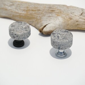 May include: Two gray stone cabinet knobs with a speckled pattern. One knob has a black base, the other a silver base. A piece of driftwood rests in the background on a white surface.