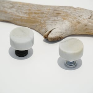 May include: Two white marble cabinet knobs with a cylindrical design, one with a black base and the other with a silver base. A piece of driftwood is in the background. The knobs are suitable for furniture.
