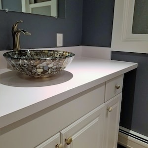 Peut inclure: Une vasque de salle de bain blanche avec un plan de travail blanc et un lavabo en verre &agrave; motif de pierre. La vasque a deux tiroirs et deux portes avec des boutons ronds dor&eacute;s.