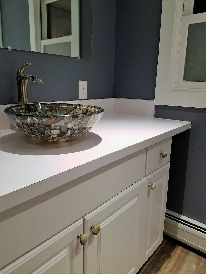 Peut inclure: Une vasque de salle de bain blanche avec un plan de travail blanc et un lavabo en verre &agrave; motif de pierre. La vasque a deux tiroirs et deux portes avec des boutons ronds dor&eacute;s.
