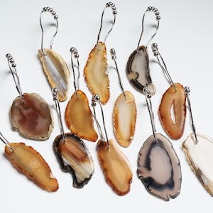 May include: A collection of agate slice shower curtain hooks. Each hook features a unique, polished agate slice in shades of brown, tan, and white, attached to a silver-toned metal hook. The hooks are arranged on a white background.