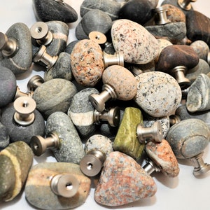 May include: A collection of smooth, natural stone drawer pulls with silver metal hardware. The stones are various shapes and colors, including gray, brown, green, and white.