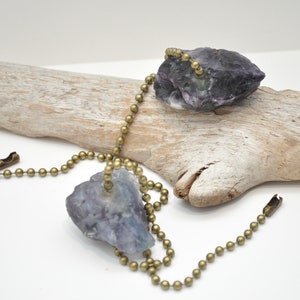 May include: Two purple fluorite crystals attached to a brass chain. The chain is looped over a piece of driftwood.