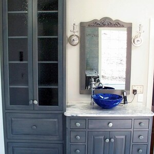 Peut inclure: Une vasque de salle de bain grise avec un plan de travail blanc et un lavabo en verre bleu. La vasque a une armoire avec des tiroirs et des portes, et une armoire haute avec des portes en verre. La vasque est dans une salle de bain avec un mur blanc et un miroir.