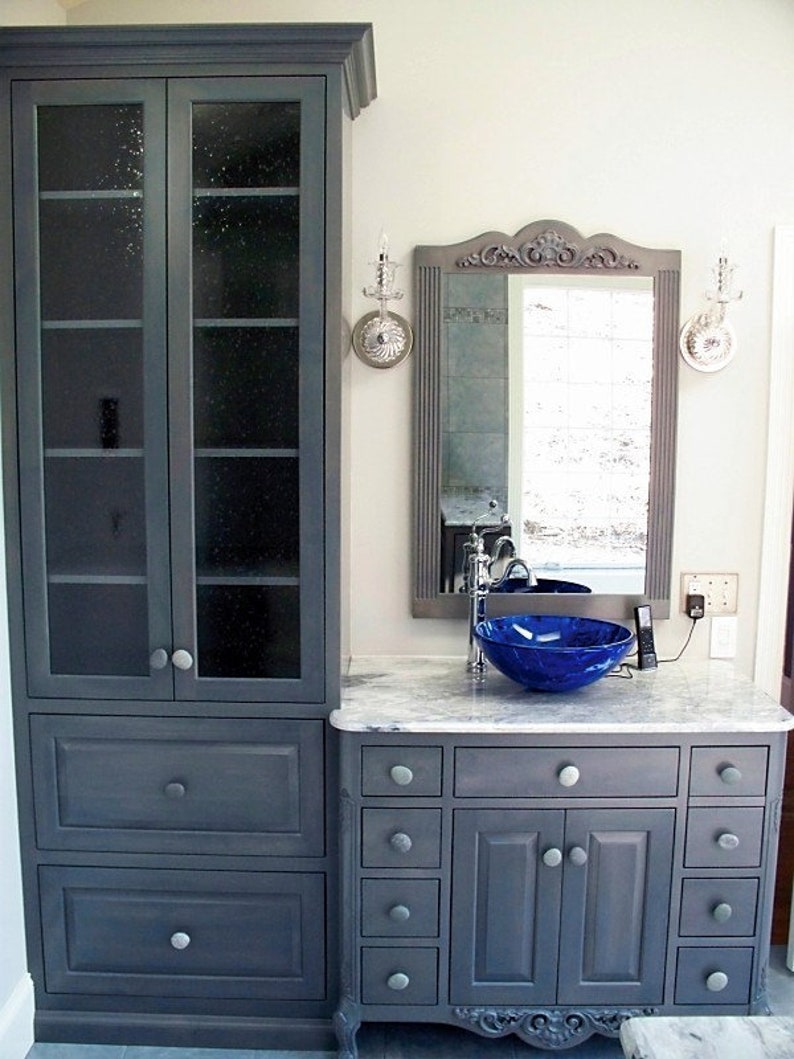 Peut inclure: Une vasque de salle de bain grise avec un plan de travail blanc et un lavabo en verre bleu. La vasque a une armoire avec des tiroirs et des portes, et une armoire haute avec des portes en verre. La vasque est dans une salle de bain avec un mur blanc et un miroir.