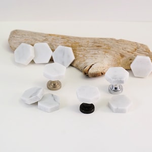 May include: White marble hexagon cabinet knobs with different colored metal bases. The knobs are arranged on a white surface with a piece of driftwood in the background.