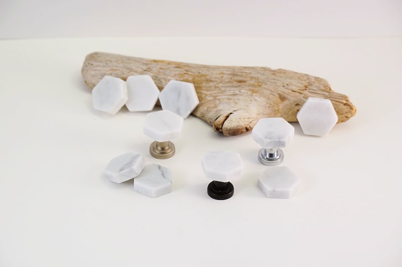 May include: White marble hexagon cabinet knobs with different colored metal bases. The knobs are arranged on a white surface with a piece of driftwood in the background.