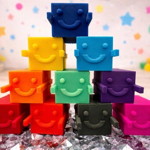 May include: A pyramid of colorful, smiling block toys in shades of red, orange, yellow, green, blue, purple, black, and pink. Each block has a smiling face and small arms. The background features a colorful star and balloon design.