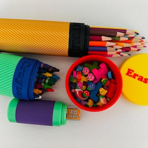 May include: A collection of school supplies including colored pencils, crayons, erasers, and pencil leads. The pencils are in a yellow tube, crayons in a blue and green container, erasers in a red container with a yellow lid that says "Erasers".