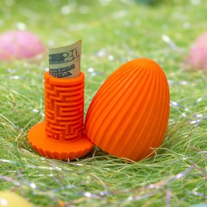 May include: An orange Easter egg-shaped container with a matching cylindrical holder. A twenty-dollar bill is partially visible in the holder. The objects are on a bed of green decorative grass with colorful Easter eggs.
