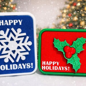 May include: Two rectangular holiday decorations. One is blue with a white snowflake and the text "HAPPY HOLIDAYS!". The other is red with a green holly design and the text "HAPPY HOLIDAYS!".