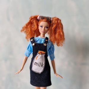 May include: A red-haired doll wearing a blue shirt and a denim overall dress with a milk carton graphic. The doll is wearing sunglasses and has her hair in pigtails.
