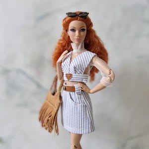 May include: A red-haired doll wearing a blue and white striped skirt, a white top with a mesh overlay, and a brown fringed bag. The doll is wearing black sunglasses and has a neutral expression.