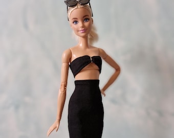 May include: A blonde doll wearing a black strapless top and a black skirt. The doll is wearing black sunglasses and has a neutral expression.