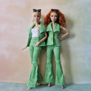May include: Two dolls are styled in matching green striped suits with flared trousers and sunglasses. One doll wears a white top, the other a green top. They stand against a soft, colourful backdrop.