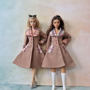 May include: Two fashion dolls wearing tan trench coats with plaid lapels and pocket flaps, red buttons, and black sunglasses perched on their heads. One doll has long blonde hair and silver heels, while the other has long brown hair and white boots.