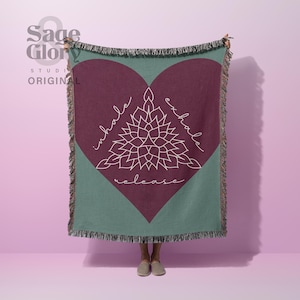May include: A woven throw blanket featuring a large dark maroon heart shape on a sage green background. The center displays a white line-art mandala design with the words inhale, exhale, and release written in a cursive script around the graphic.